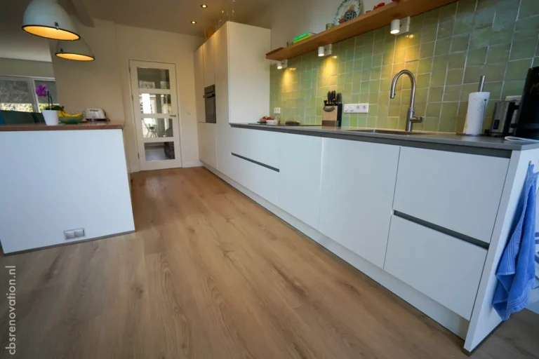 Modern kitchen renovation featuring sleek white cabinetry, a grey countertop, and green tiled backsplash, with wooden flooring.