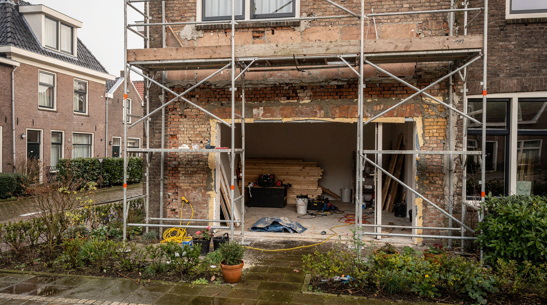 A Dutch row house renovation with scaffolding, open brickwork, a contractor’s tools inside, and a small garden under soft overcast daylight.